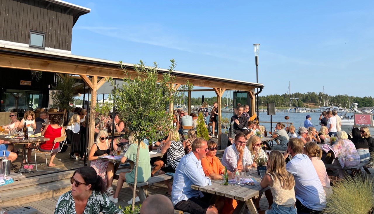 Mänsikor som äter vid bord på restaurangen Kölsvinets uteservering. I bakgrunden står några och spelar musik.