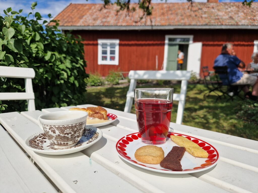 En kaffekopp, ett glas röd saft och två assietter med fika på. I bakgrunden ett rött torp.