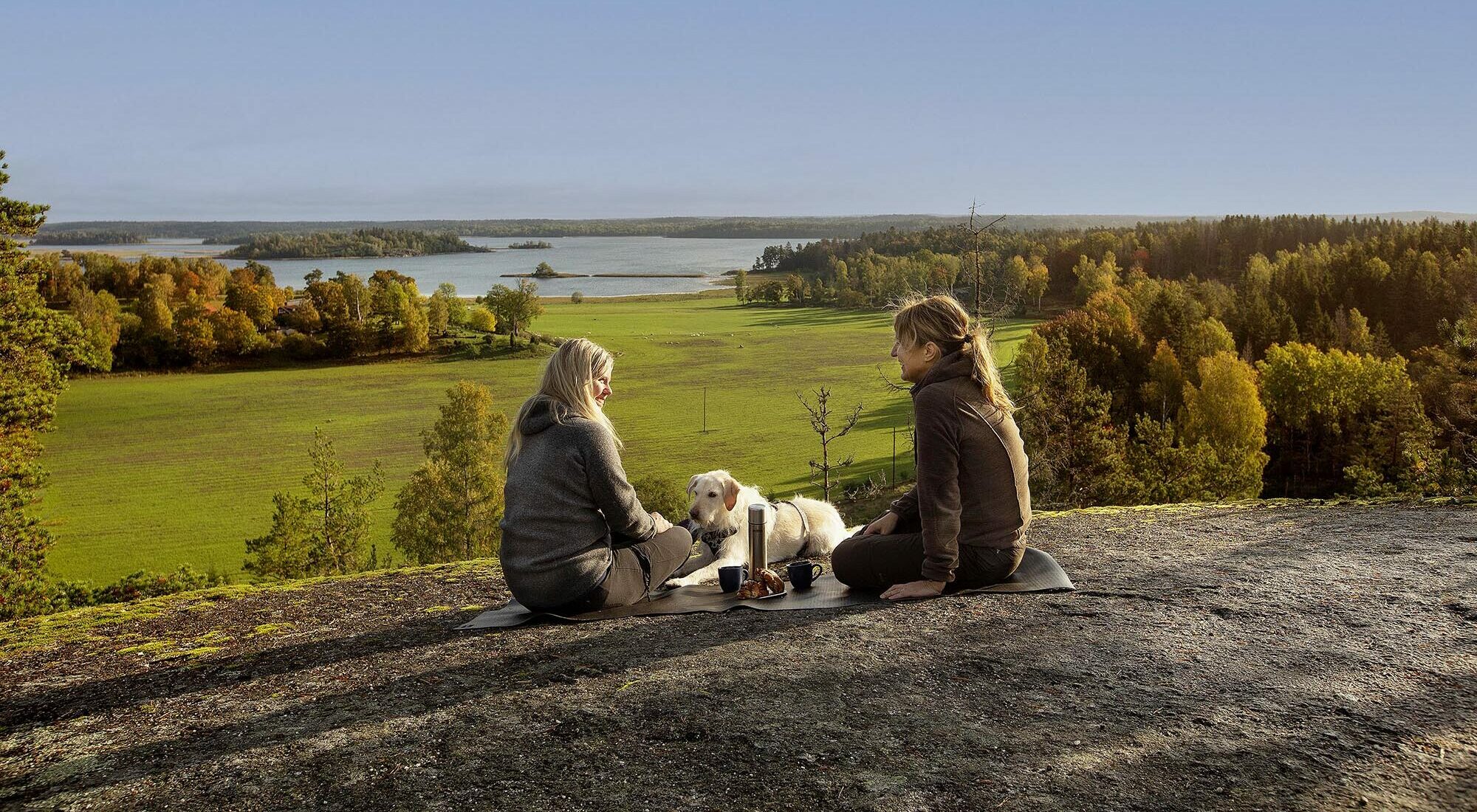 Två kvinnor och en hund sitter på berg och fikar